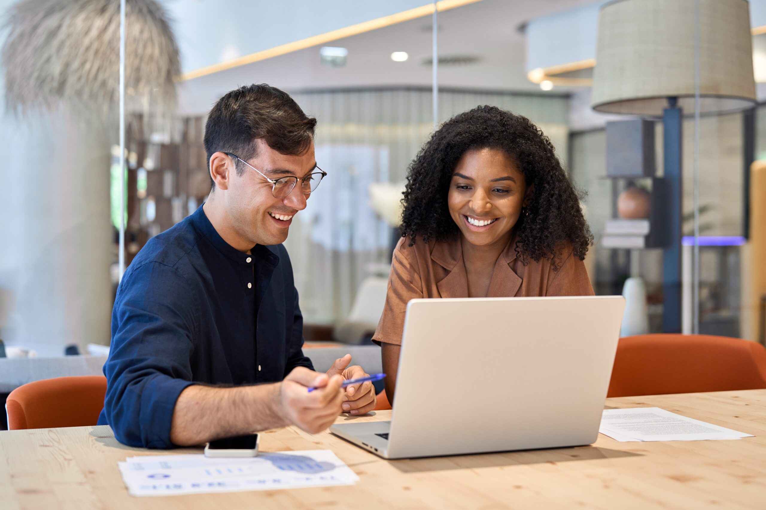 Two people sit at a table in a modern office, smiling and looking at a laptop screen, with papers and a pen on the table.