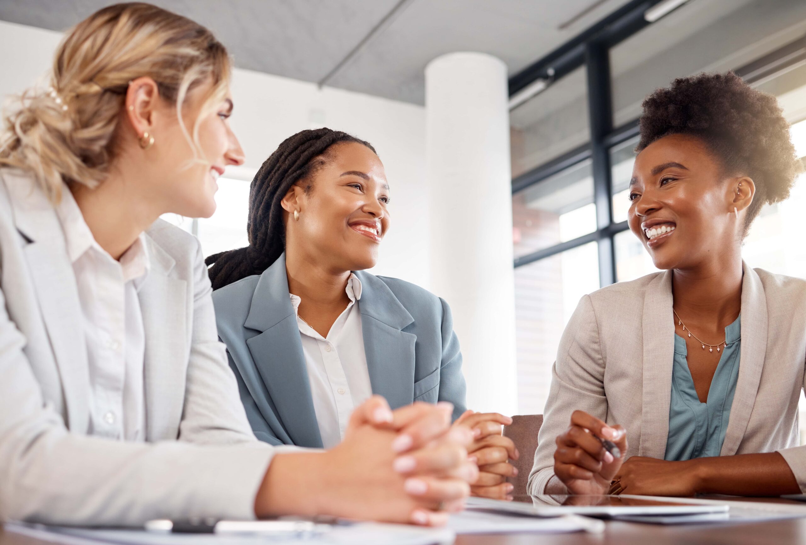 Three women in business attire sit at a table, smiling and talking, in a modern office setting with natural light.